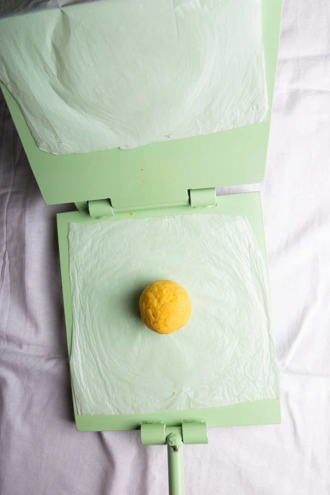 A small yellow dough ball sits on the center of an open green tortilla press lined with parchment paper, ready to make corn tortillas on a white fabric surface. The press is set for flattening the dough and crafting homemade corn tortillas.