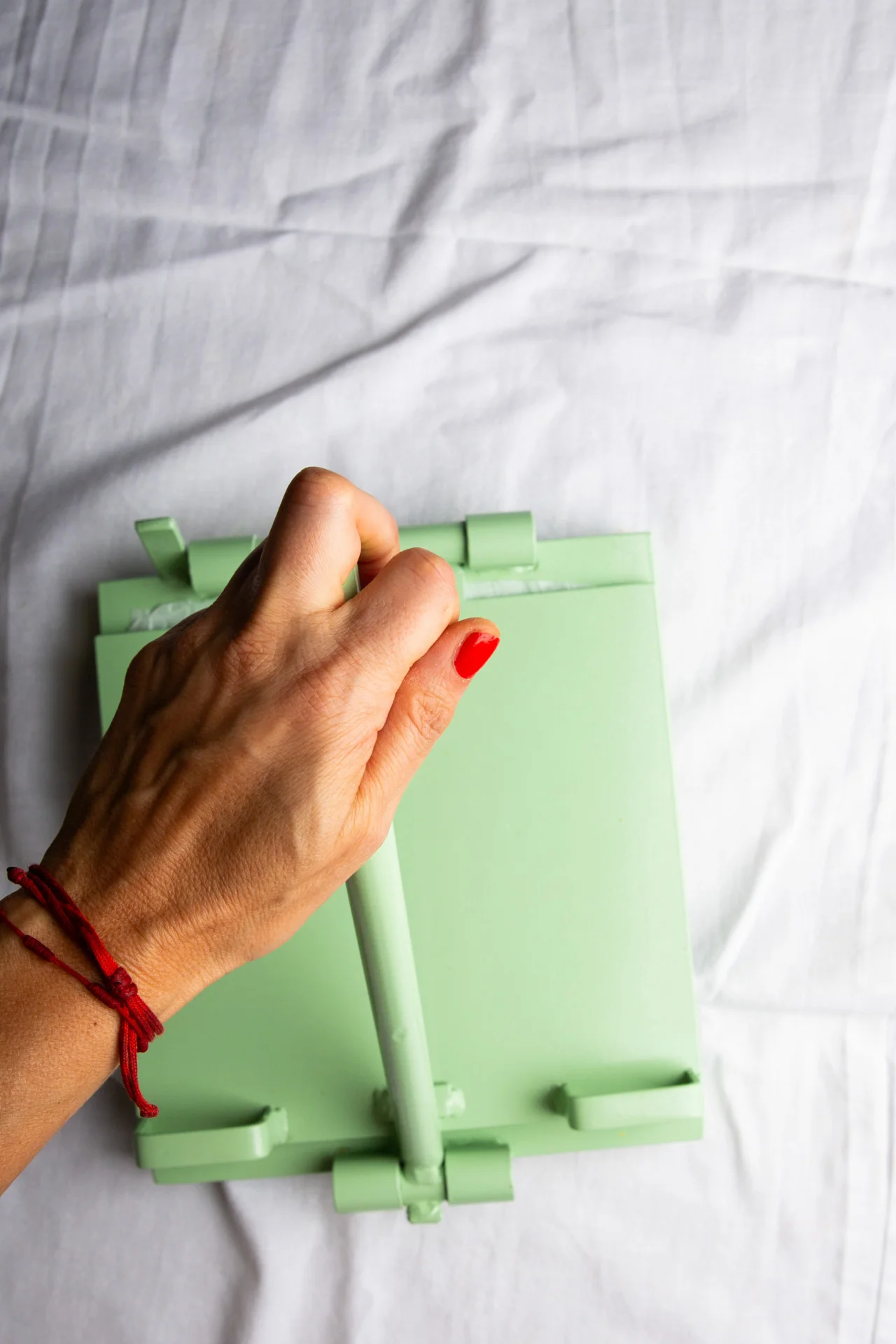 A hand with red nail polish and a red string bracelet grips the handle of a mint green mop against a white fabric background, evoking the care and precision used to make corn tortillas. The mop stands upright in bright lighting.