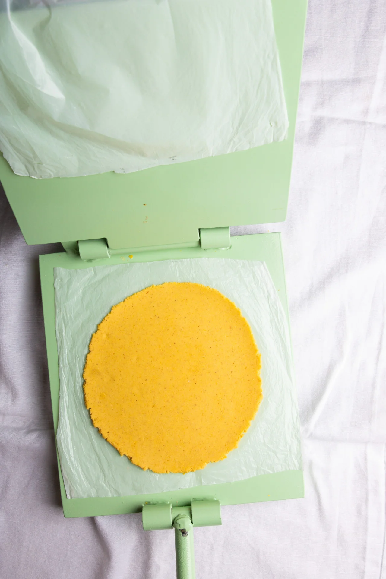A round, flat yellow disc of dough sits on parchment paper inside a light green tortilla press, ready to make corn tortillas. The neatly pressed edges and parchment lining ensure perfectly shaped homemade corn tortillas every time.
