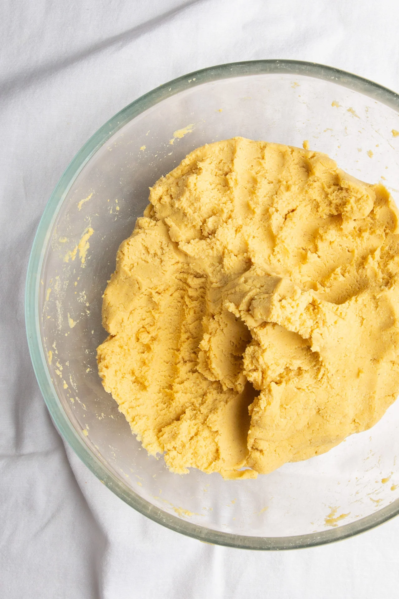 A large, clear glass bowl containing a mound of pale yellow dough sits on a white cloth surface. The dough appears soft and slightly crumbly, with visible ridges—perfect for those who want to make corn tortillas from scratch.
