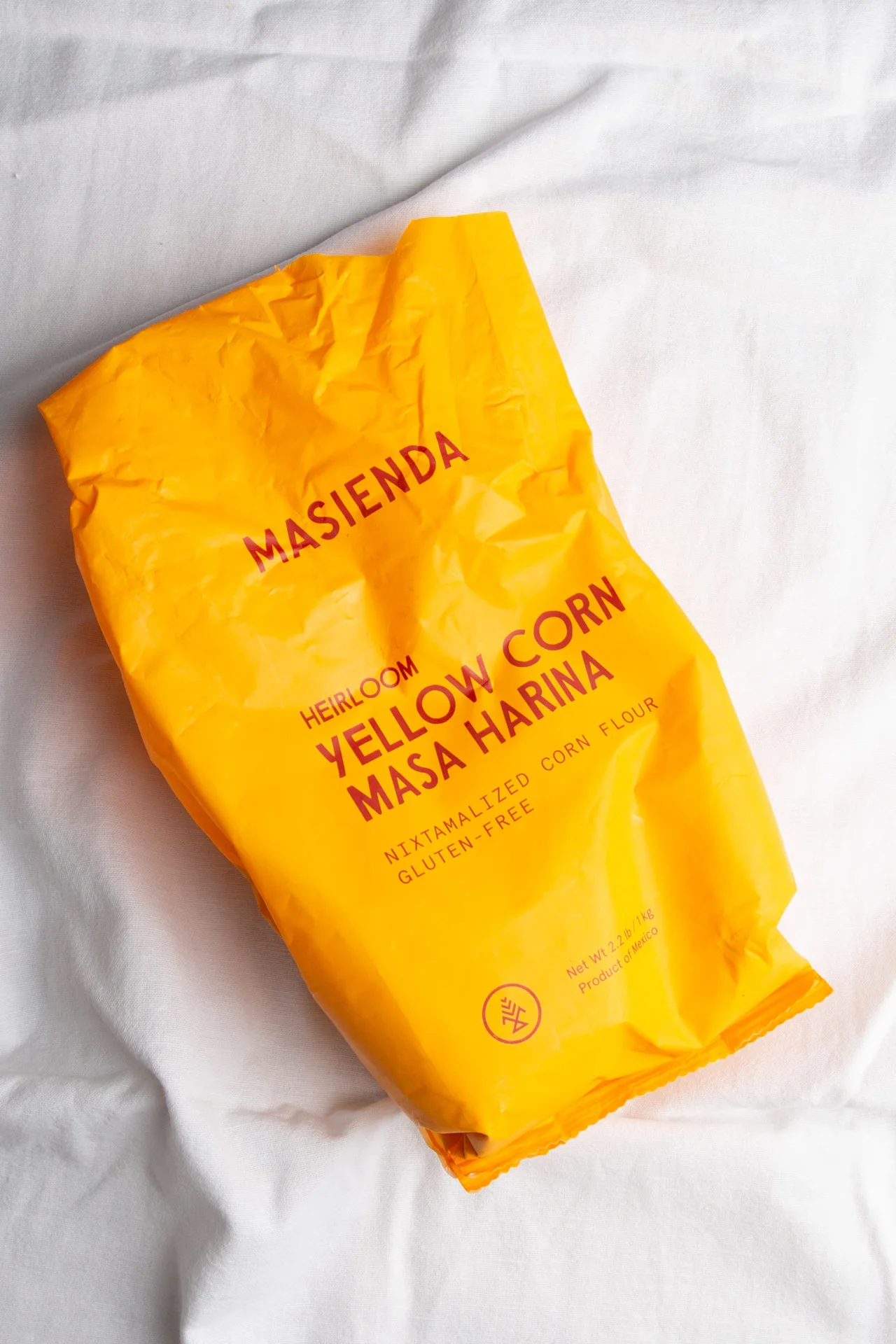 A crumpled, bright yellow bag of Masienda Heirloom Yellow Corn Masa Harina lies on a white fabric surface. Perfect to make corn tortillas, the packaging states it is nixtamalized corn flour and gluten-free, with text in red and a small round logo.