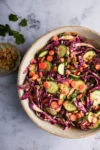 Sobre una superficie de mármol descansa un cuenco de ensalada de colores con rodajas de pepino, col lombarda, zanahorias, cilantro y cacahuetes. A la izquierda, un platito con cacahuetes y totopos crujientes aporta textura crujiente y un toque de color a esta fresca presentación.