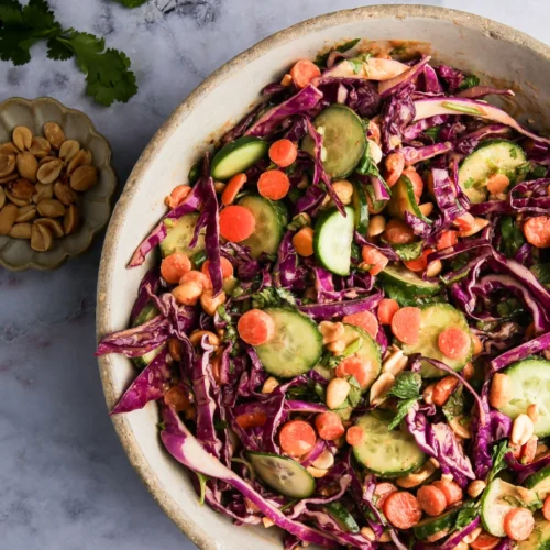 Sobre una superficie de mármol descansa un cuenco de ensalada de colores con rodajas de pepino, col lombarda, zanahorias, cilantro y cacahuetes. A la izquierda, un platito con cacahuetes y totopos crujientes aporta textura crujiente y un toque de color a esta fresca presentación.