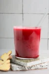 Un vaso transparente lleno de jugo de remolacha de color rojo intenso reposa sobre dos posavasos de terrazo apilados. En el interior del vaso hay una pajita transparente. Junto a los posavasos hay dos rodajas de jengibre fresco. El fondo presenta paredes de azulejos blancos con líneas de lechada visibles.