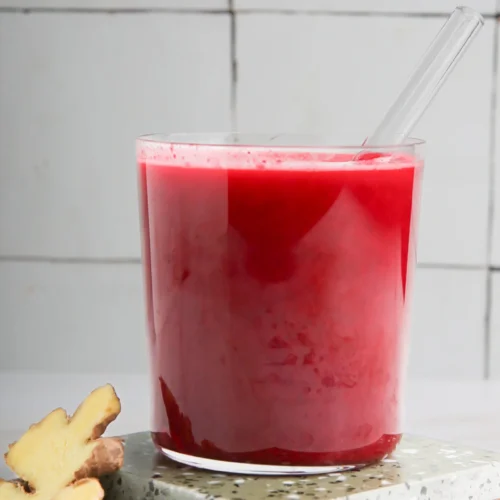 Un vaso transparente lleno de jugo de remolacha de color rojo intenso reposa sobre dos posavasos de terrazo apilados. En el interior del vaso hay una pajita transparente. Junto a los posavasos hay dos rodajas de jengibre fresco. El fondo presenta paredes de azulejos blancos con líneas de lechada visibles.