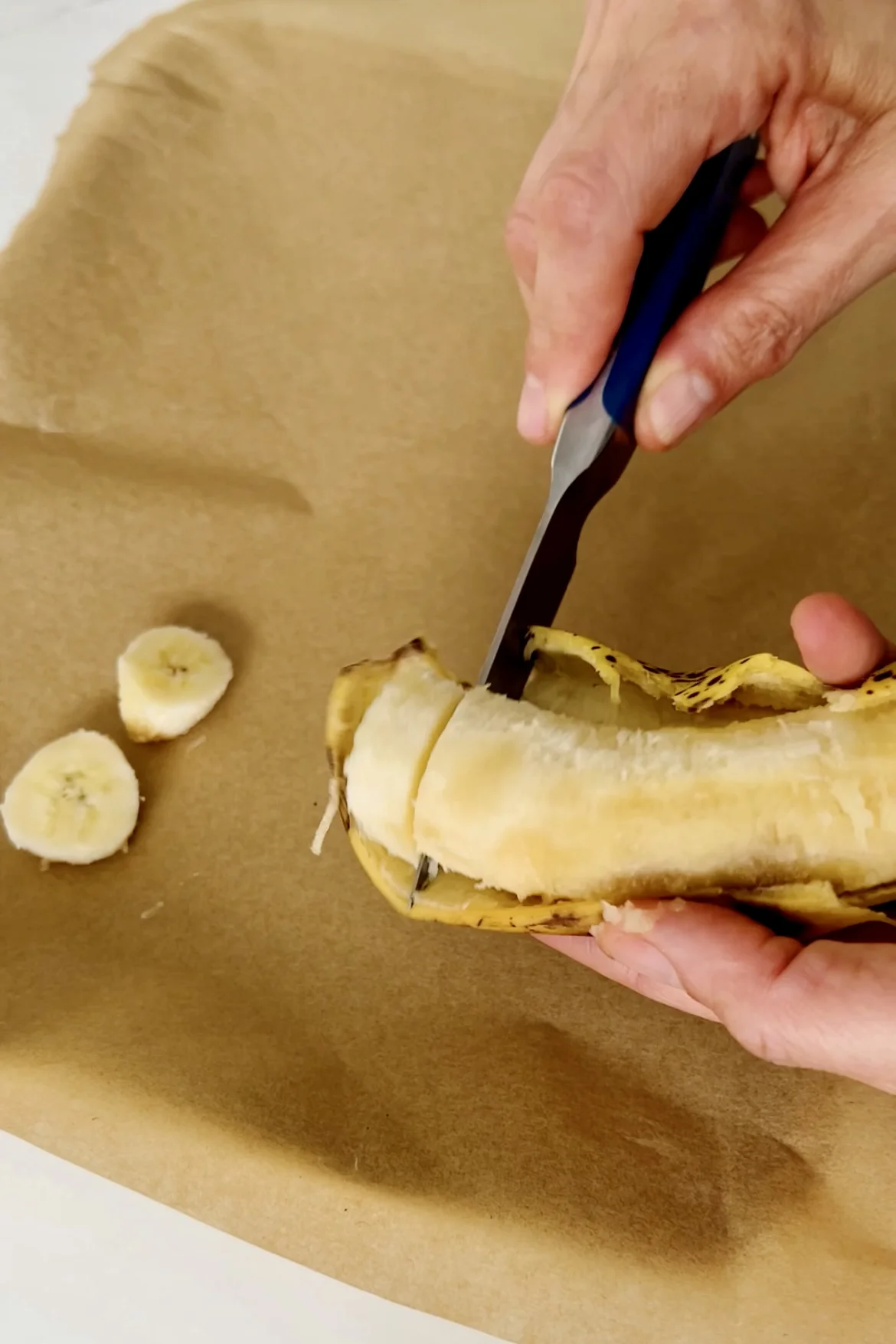 Manos pelando y cortando un plátano con un cuchillo de mango azul sobre papel de pergamino marrón. El plátano parcialmente pelado muestra una pulpa amarilla madura, mientras que la cáscara cuelga hacia abajo. Dos rodajas de plátano redondas yacen sobre el papel cercano.
