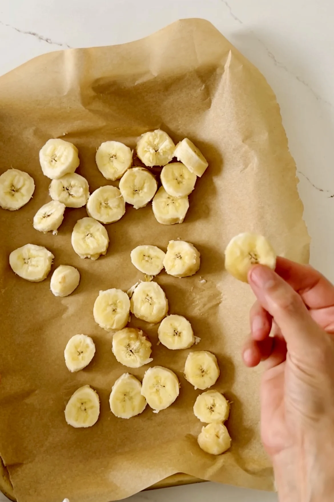 Una mano sostiene una rodaja de plátano sobre una bandeja para hornear cubierta con papel de pergamino. Varias rodajas de plátano están distribuidas uniformemente sobre la bandeja. Las bananas son de color amarillo pálido y el papel de pergamino es de color marrón claro. La superficie debajo es blanca, posiblemente una encimera.