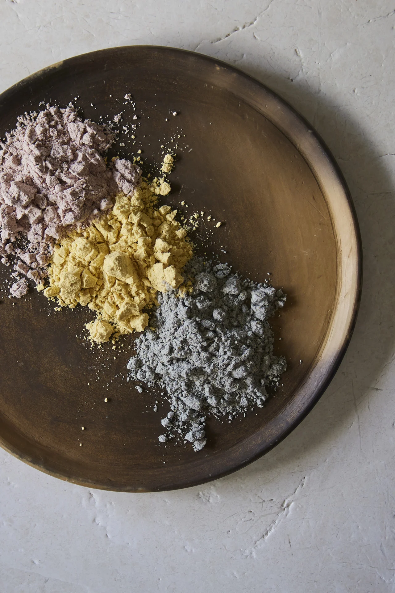 A round wooden plate holds three small piles of colored powders—perfect for making homemade corn tortillas: purple on the upper left, yellow in the center, and gray on the lower right. The plate rests on a light, textured surface.
