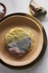 A stack of perfect tri-colored corn tortillas—yellow, pink, and blue—sits on a tan ceramic plate atop a brown saucer. Torn pieces rest nearby, with a wooden bowl of coarse salt on the textured white surface for an inviting make at home treat.