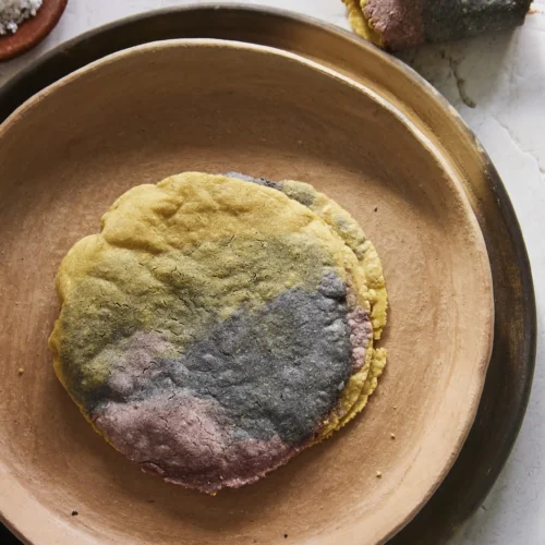A stack of perfect tri-colored corn tortillas—yellow, pink, and blue—sits on a tan ceramic plate atop a brown saucer. Torn pieces rest nearby, with a wooden bowl of coarse salt on the textured white surface for an inviting make at home treat.