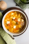 Un cuenco de curry colorido presenta cubos de paneer y papas cortadas en cubitos en un rico caldo de color naranja, aderezado con hierbas. El cuenco reposa sobre un plato de madera con una servilleta verde y una cuchara a un lado. Un salero de cerámica y una estera verde con textura se encuentran en el fondo.