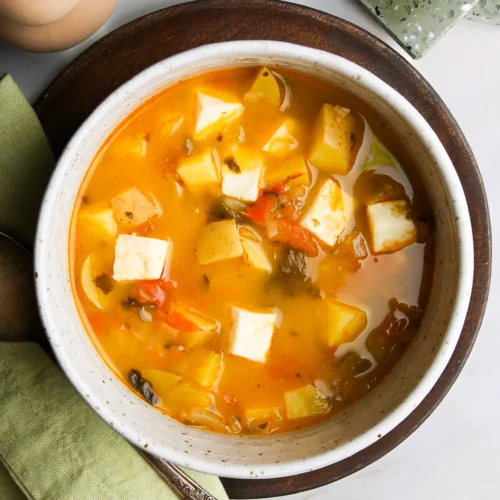 Un cuenco de curry colorido presenta cubos de paneer y papas cortadas en cubitos en un rico caldo de color naranja, aderezado con hierbas. El cuenco reposa sobre un plato de madera con una servilleta verde y una cuchara a un lado. Un salero de cerámica y una estera verde con textura se encuentran en el fondo.