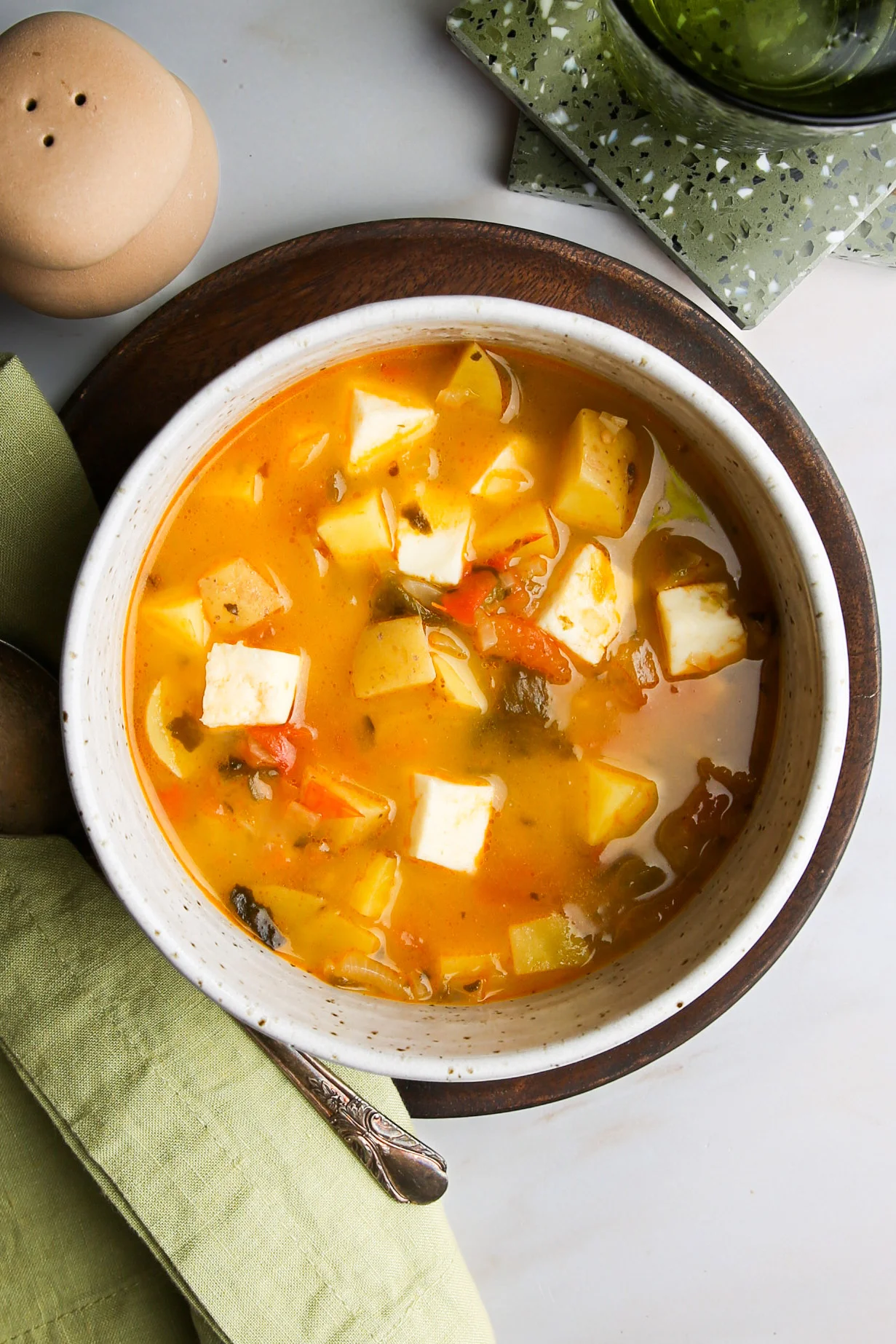 A bowl of vegetable curry soup inspired by Sonoran Mexican Potato Soup with diced paneer, potatoes, tomatoes, and herbs in a yellow-orange broth sits on a wooden plate. A green napkin, silver spoon, coaster, and tan pepper shaker are nearby.