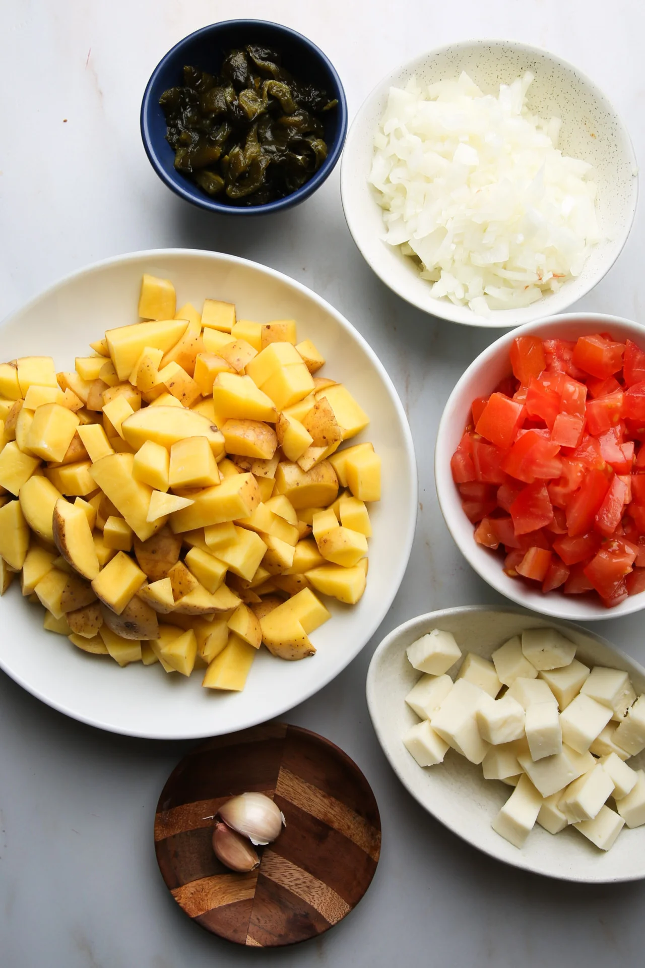 Chopped ingredients for Caldo de Queso—Sonoran Mexican Potato Soup—include diced potatoes on a white plate, onions and tomatoes in bowls, sliced mozzarella cheese, chopped green chili in a blue bowl, and garlic cloves on a wooden dish.