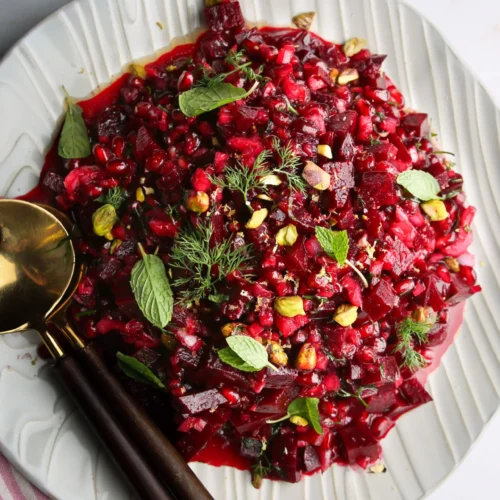 En un plato blanco se sirve una ensalada vibrante de remolacha y granada. La ensalada está adornada con hojas de menta fresca y eneldo, y espolvoreada con pistachos para darle más textura. Un par de cucharas para servir con mango oscuro descansan en el borde del plato, sobre una superficie clara con un paño de rayas cerca.