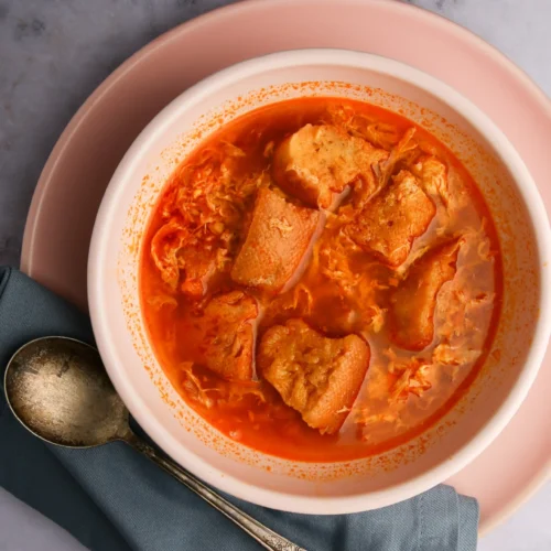 Un cuenco de sopa de tomate cubierto con rebanadas de pan, colocado sobre un plato de color salmón. Una cuchara de plata antigua reposa sobre un paño verde azulado doblado junto al cuenco. El fondo es una superficie de mármol gris claro, que le da una presentación elegante y acogedora.