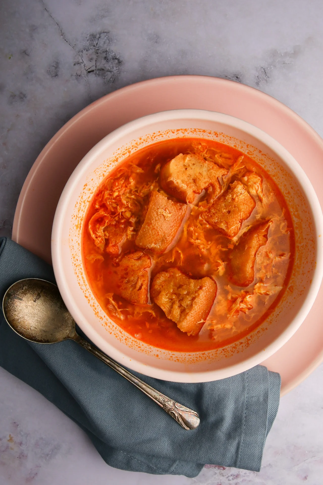 Plato hondo con sopa de ajo y trozos de pan flotando, servido sobre un plato rosa con una cuchara de plata vintage y una servilleta verde azulado.