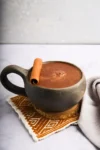 Una taza de chocolate caliente coronada con una rama de canela reposa sobre un posavasos tejido. La taza es oscura y redondeada, con un asa robusta. Debajo hay un paño con estampados blancos y naranjas. El fondo es una superficie de mármol gris claro y suave.