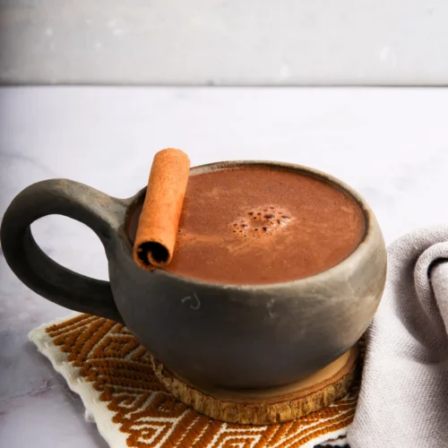 Una taza de chocolate caliente coronada con una rama de canela reposa sobre un posavasos tejido. La taza es oscura y redondeada, con un asa robusta. Debajo hay un paño con estampados blancos y naranjas. El fondo es una superficie de mármol gris claro y suave.
