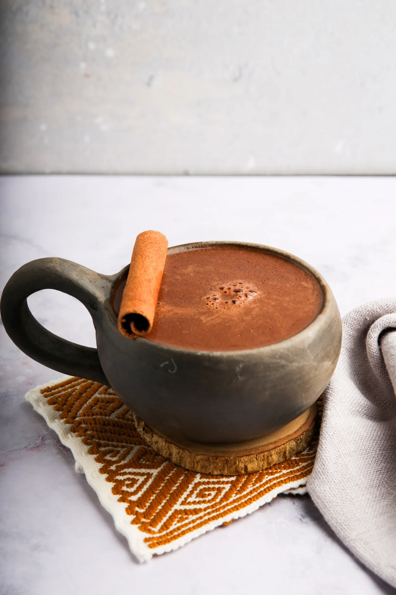 Una taza de chocolate caliente coronada con una rama de canela reposa sobre un posavasos tejido. La taza es oscura y redondeada, con un asa robusta. Debajo hay un paño con estampados blancos y naranjas. El fondo es una superficie de mármol gris claro y suave.