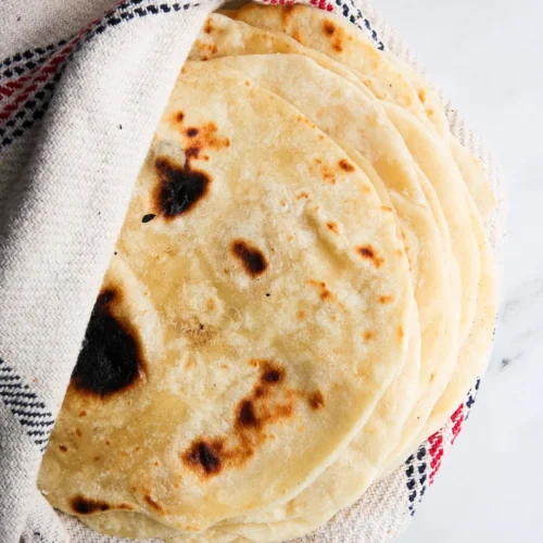 Sobre una superficie de mármol se encuentra una pila de panes planos redondos de color marrón dorado envueltos parcialmente en un paño blanco con rayas rojas y negras. Los panes planos tienen manchas oscuras y de cocción irregular, lo que indica que se calentaron en una sartén o plancha.