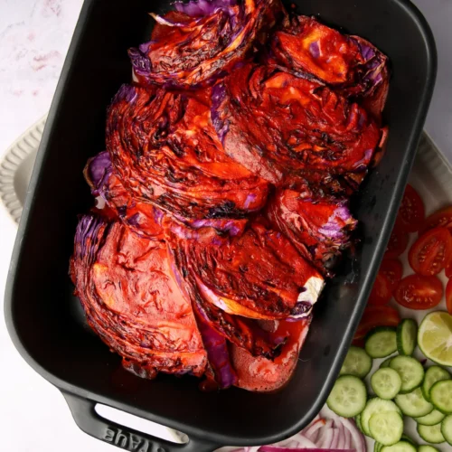 En una fuente para horno negra se colocan rodajas de col a la parrilla bañadas en una marinada roja. Al lado, hay rodajas de pepino, aros de cebolla roja y rodajas de tomate, junto con una rodaja de lima en una bandeja blanca. El fondo es una superficie clara y jaspeada.