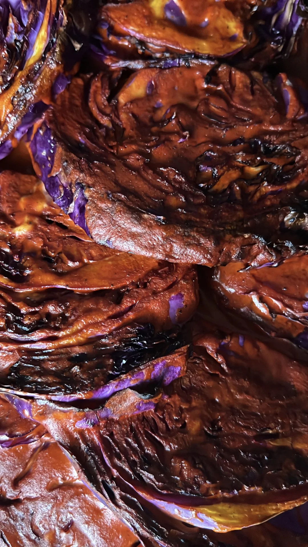 Primer plano de rebanadas de col asada con bordes bien dorados y tonos intensos entre morado y caf&eacute; rojizo, mostrando distintas texturas: partes crujientes y caramelizadas junto a secciones suaves y brillosas. Las capas superpuestas crean un patr&oacute;n visual rico y lleno de detalle.