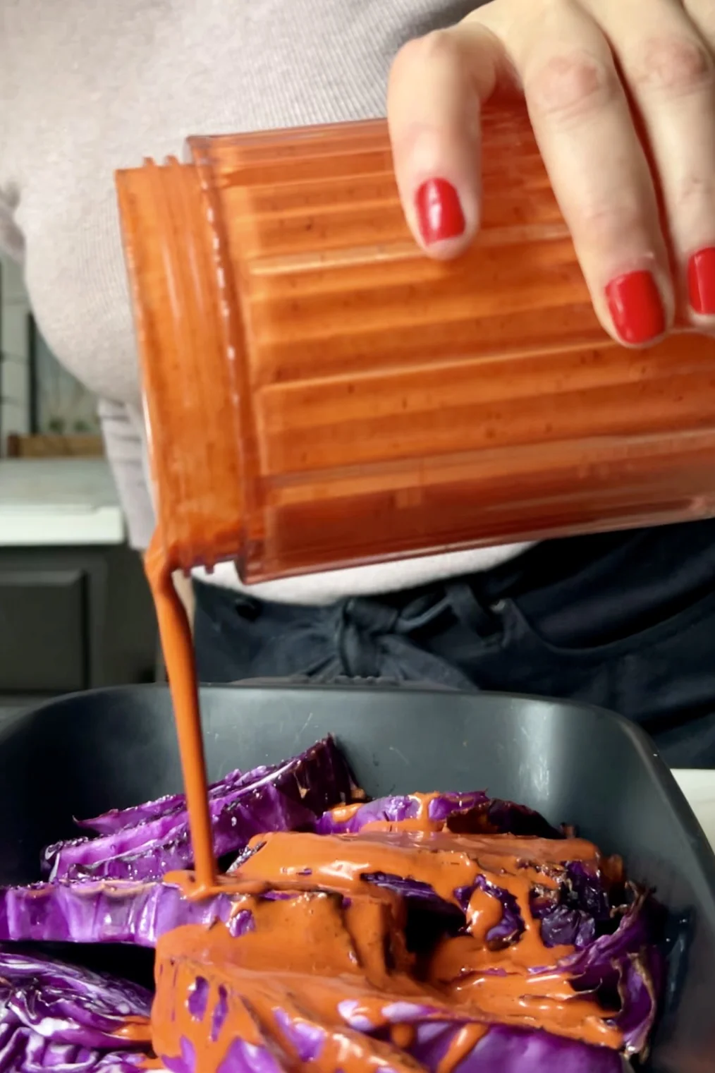 Una persona con esmalte de u&ntilde;as rojo vierte una salsa zarandeada espesa de color naranja desde un recipiente acanalado sobre col morada picada en una charola negra. La persona lleva una blusa clara y pantalones oscuros, con una cocina visible de fondo.