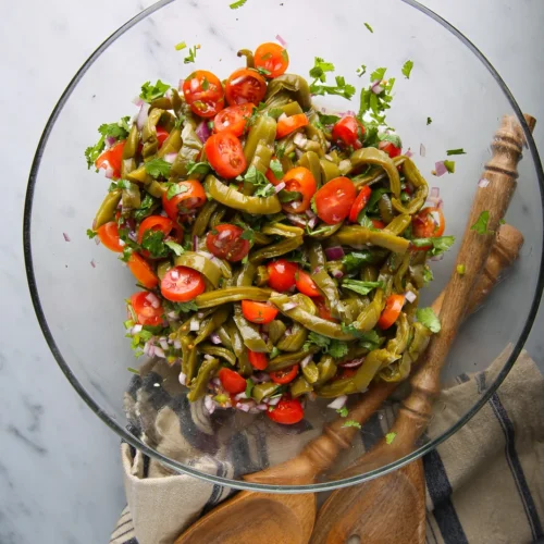 Cuenco de cristal lleno de ensalada de judías verdes, tomates cherry, cebolla morada y cilantro, sobre una superficie de mármol gris. Los utensilios de madera para ensalada descansan sobre un paño rayado debajo del cuenco. La ensalada se ve fresca y vibrante con una mezcla de colores verde, rojo y morado.