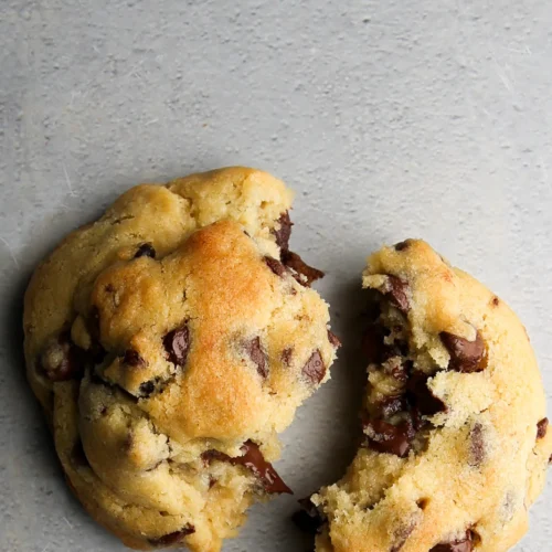 Dos galletas con chispas de chocolate sobre una superficie gris. Una galleta está intacta, mientras que la otra está partida por la mitad, revelando trozos de chocolate derretido en el interior. Las galletas son suaves y de color marrón dorado, salpicadas de trozos de chocolate visibles, lo que muestra un aspecto recién horneado.