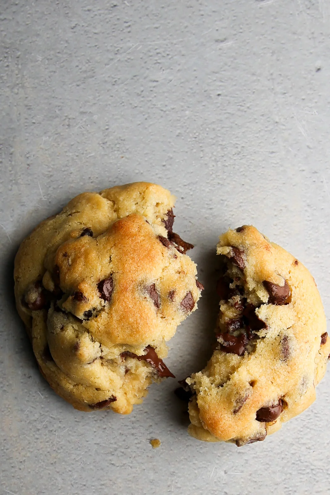 Dos galletas estilo Levain Bakery con chispas de chocolate sobre una superficie gris, una partida a la mitad mostrando el centro suave y derretido con trozos de chocolate.