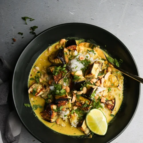 A dark, round bowl filled with creamy yellow curry featuring chunks of grilled eggplant, rice, and topped with chopped peanuts and cilantro. A lime wedge sits on the side of the bowl. A striped cloth napkin is partially visible on the left, set against a textured gray background.