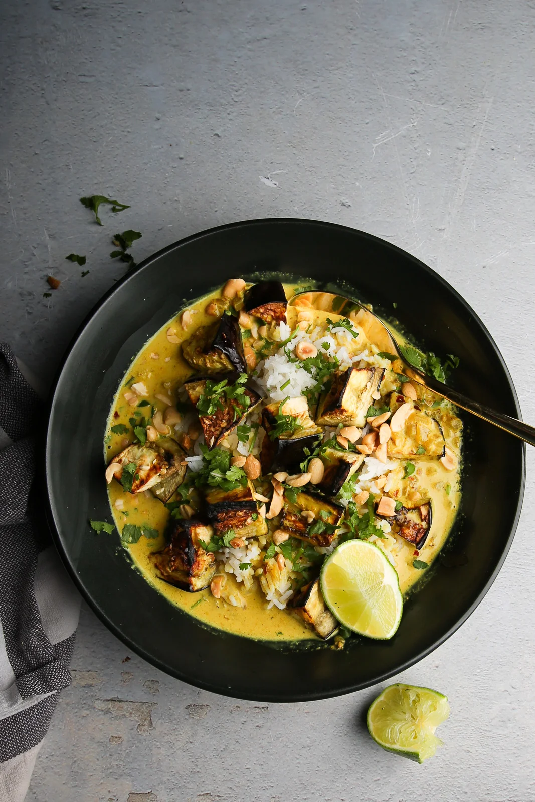 A dark, round bowl filled with creamy yellow curry featuring chunks of grilled eggplant, rice, and topped with chopped peanuts and cilantro. A lime wedge sits on the side of the bowl. A striped cloth napkin is partially visible on the left, set against a textured gray background.