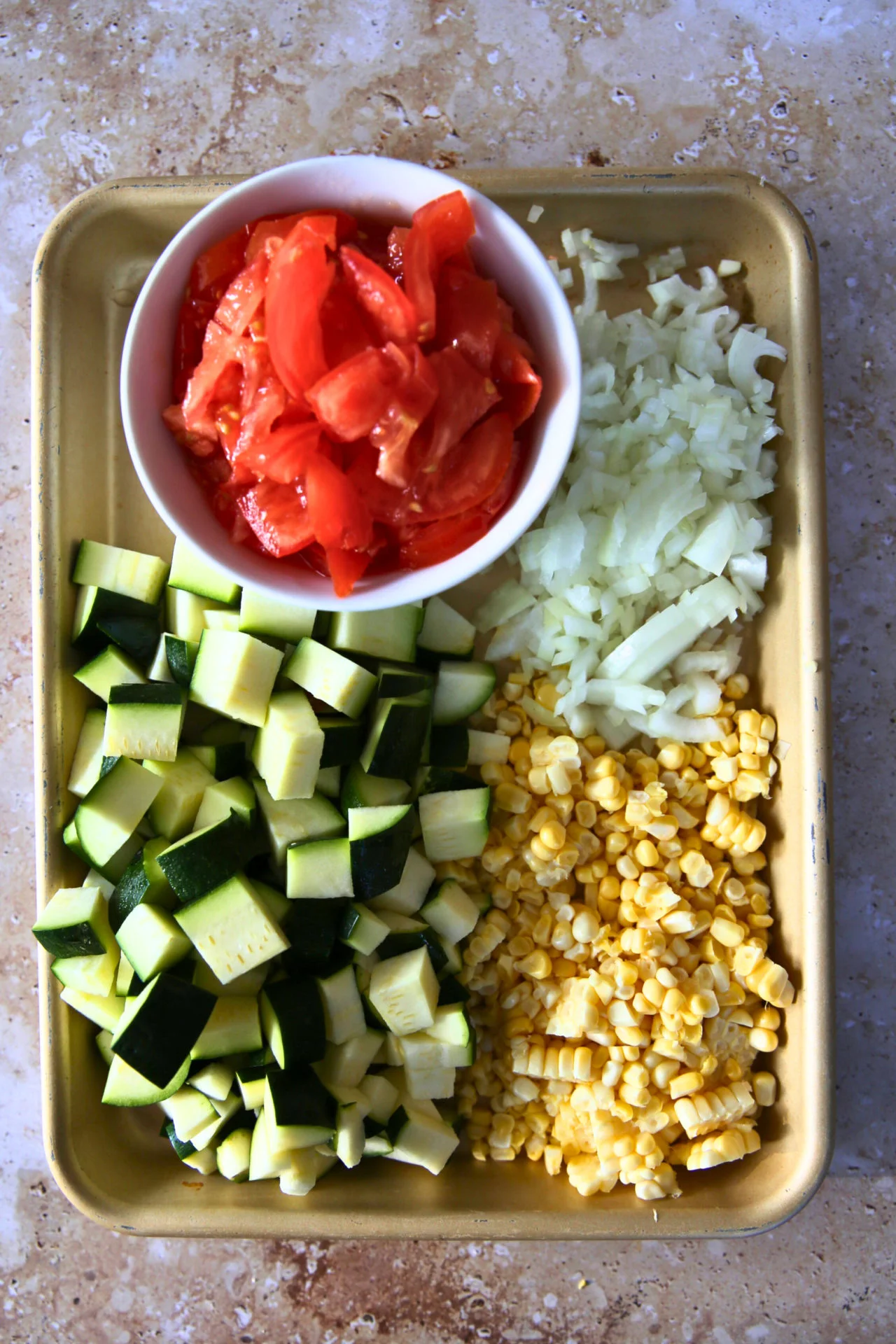 Ingredientes para calabacitas con queso sobre una bandeja: calabacitas picadas, granos de elote, cebolla picada y un tazón de tomate