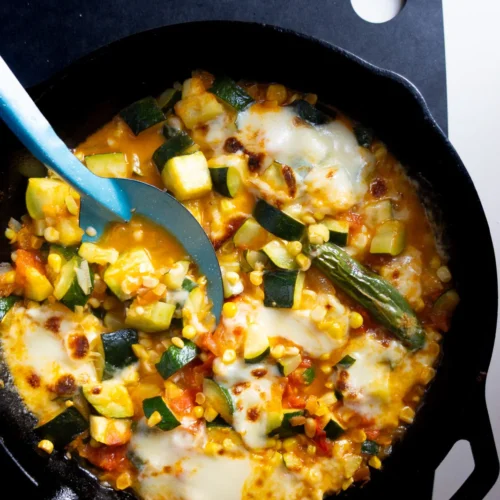 Una sartén de hierro fundido llena de un colorido plato de verduras con calabacín, maíz, tomate y chiles verdes. Los ingredientes se mezclan con queso derretido, creando una textura cremosa. Una cuchara azul reposa en la sartén, sobre una tabla de cortar negra con un agujero redondo.