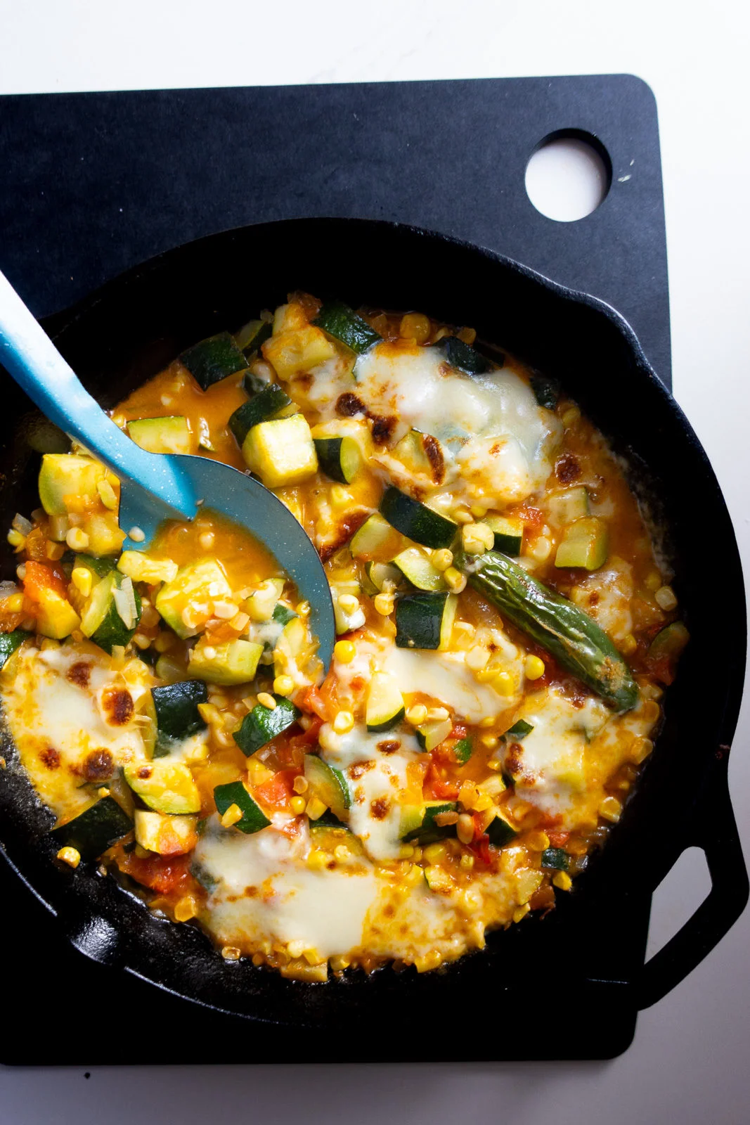 Una sart&eacute;n de hierro fundido llena de un colorido plato de verduras con calabac&iacute;n, ma&iacute;z, tomate y chiles verdes. Los ingredientes se mezclan con queso derretido, creando una textura cremosa. Una cuchara azul reposa en la sart&eacute;n, sobre una tabla de cortar negra con un agujero redondo.