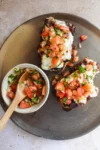 A ceramic plate holds two Molletes topped with melted cheese and fresh salsa, including tomatoes, onions, and herbs. Beside the bread, a small bowl contains extra salsa with a wooden spoon, all set on a light stone surface—an authentic touch of Mexican food delight.