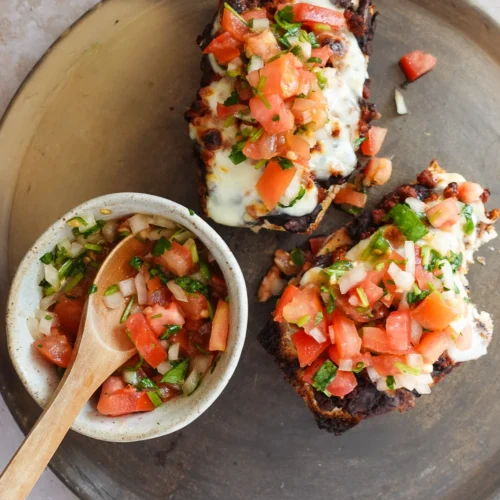 Vista de arriba hacia abajo de dos rebanadas de pan tostado cubiertas con queso derretido, frijoles negros y salsa fresca en un plato redondo. Un tazón pequeño de salsa con tomates cortados en cubitos, cebollas y cilantro se encuentra al lado con una cuchara de madera. El fondo tiene una superficie clara y texturizada.
