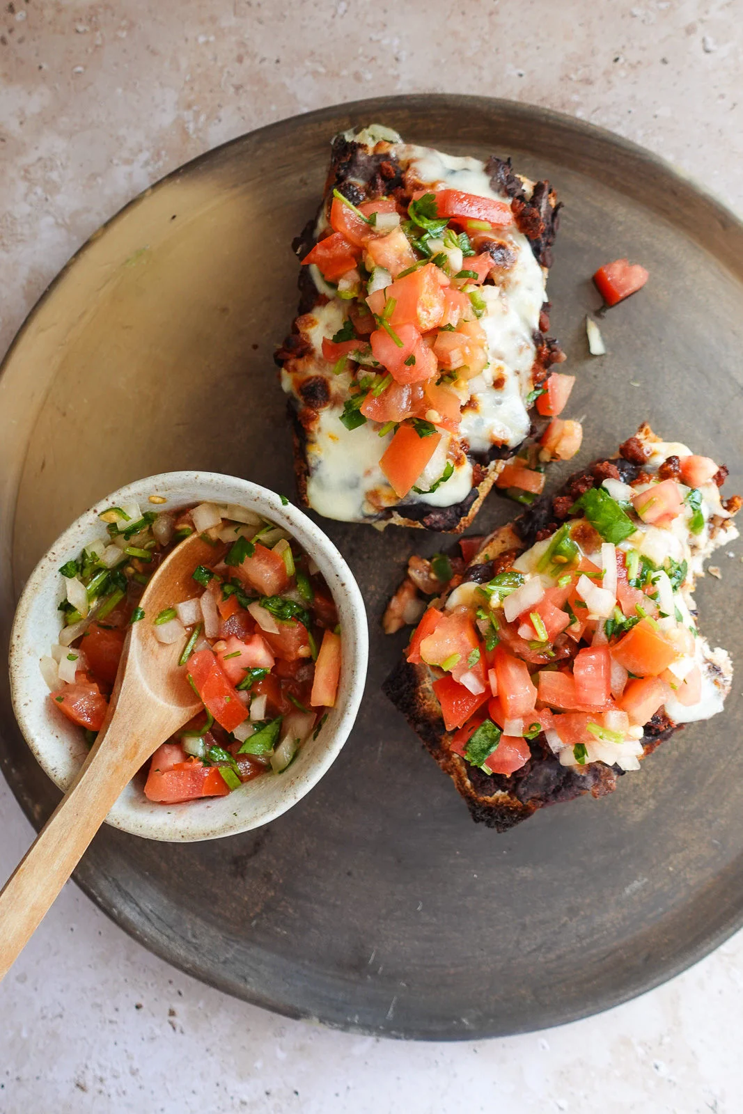Vista de arriba hacia abajo de dos rebanadas de pan tostado cubiertas con queso derretido, frijoles negros y salsa fresca en un plato redondo. Un taz&oacute;n peque&ntilde;o de salsa con tomates cortados en cubitos, cebollas y cilantro se encuentra al lado con una cuchara de madera. El fondo tiene una superficie clara y texturizada.