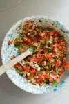 Un cuenco de cerámica con motas azules contiene salsa fresca, con tomates picados, cebollas, cilantro y jalapeños. Una cuchara de madera reposa en el cuenco, mezclando los coloridos ingredientes, todo sobre una encimera de color gris claro.