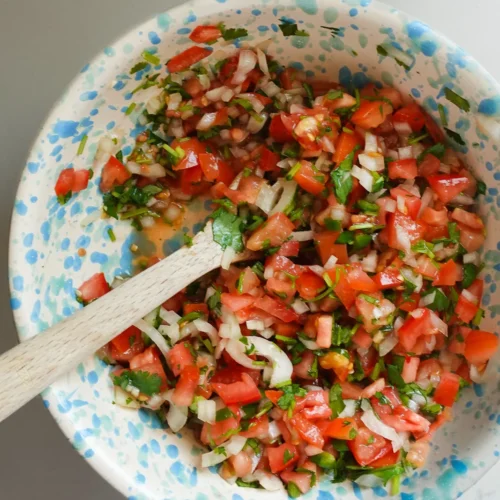 Un cuenco de cerámica con motas azules contiene salsa fresca, con tomates picados, cebollas, cilantro y jalapeños. Una cuchara de madera reposa en el cuenco, mezclando los coloridos ingredientes, todo sobre una encimera de color gris claro.