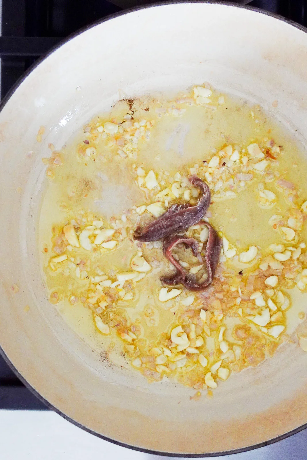 A close-up of a light-colored pan on a stove shows sizzling chopped garlic and anchovy fillets cooking in oil—perfect for a quick weeknight pasta. The garlic is golden brown, and the anchovies are melting into the oil for rich anchovy spaghetti.