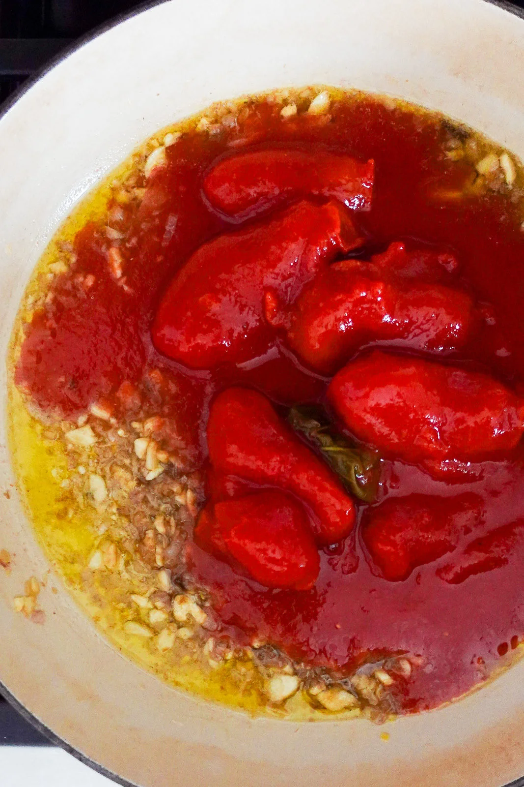 A close-up of a pot on a stove contains whole peeled tomatoes in red sauce, chopped garlic, oil, herbs, and anchovy. The ingredients are just beginning to cook and blend, perfect for starting a savory Weeknight Pasta or classic Tomato Spaghetti.