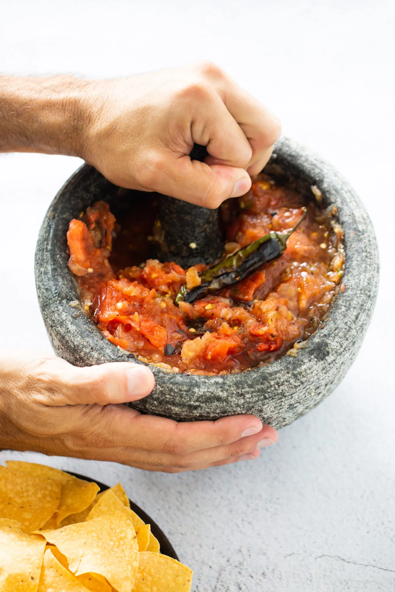 Dos manos utilizan expertamente un molcajete, el utensilio tradicional de la cocina casera mexicana, para moler tomates asados, chiles y ajo y convertirlos en una salsa roja vibrante y con trozos. En la esquina inferior izquierda, un plato negro sostiene crujientes tortillas de maíz amarillas.
