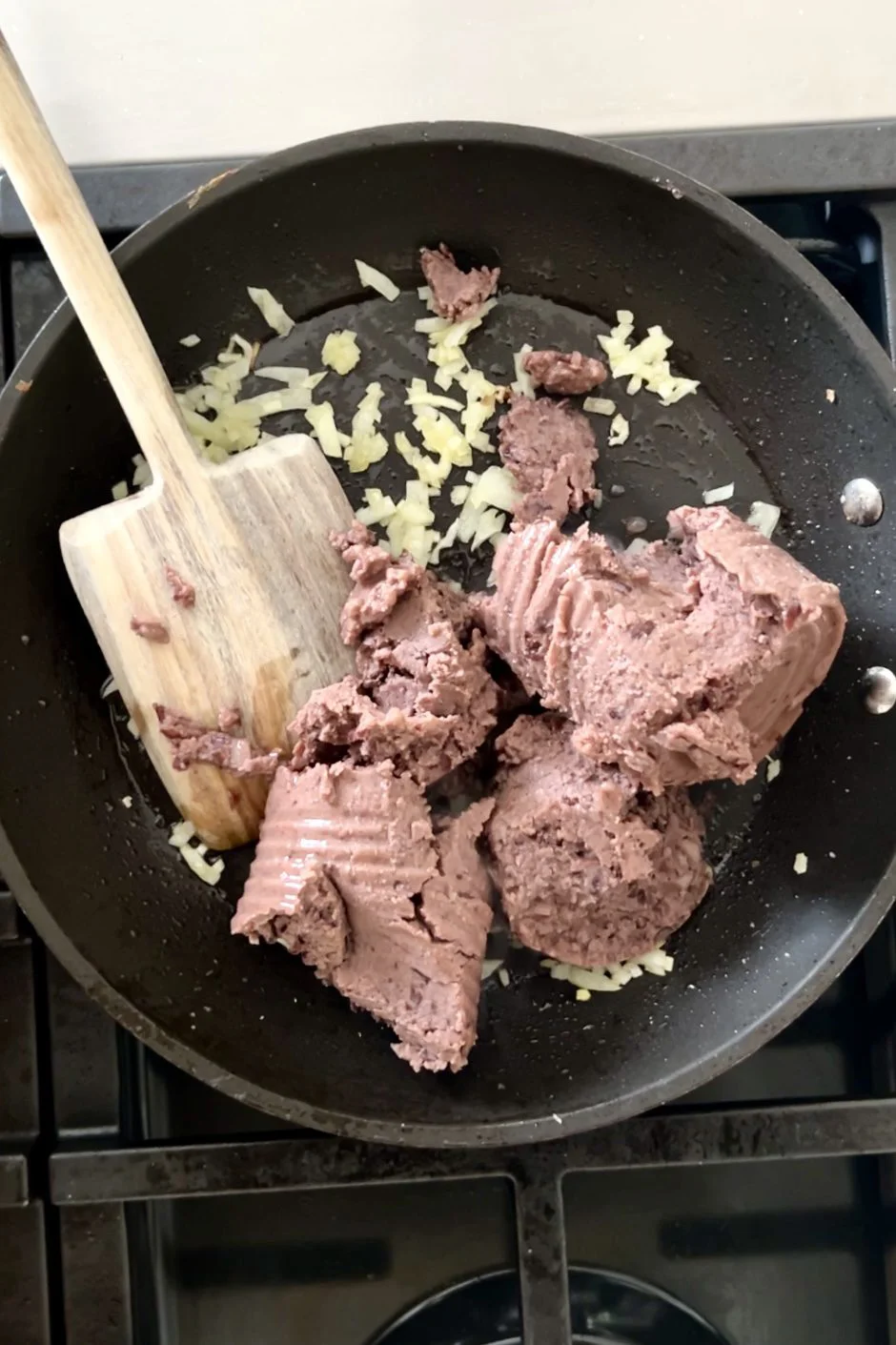 Un sartén en la estufa con cebolla picada sofriéndose junto con varias cucharadas de carne enlatada. Una espátula de madera reposa en el sartén mezclando los ingredientes, similar a como se recalientan los frijoles refritos en la estufa.