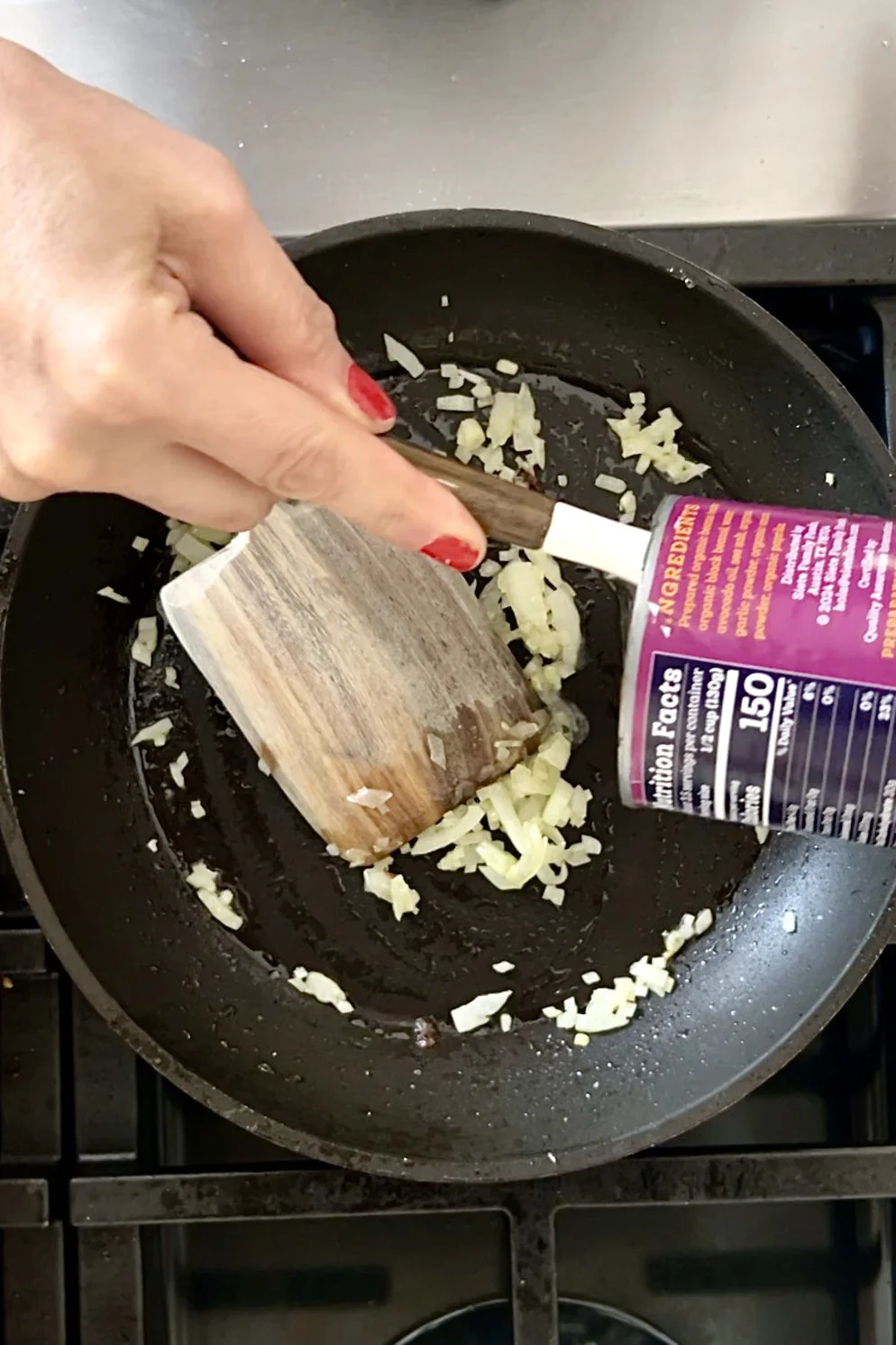 Una mano con esmalte rojo revuelve cebolla picada en un sartén negro mientras otra persona agrega sal desde un envase morado—perfecto para preparar frijoles refritos o antes de recalentar frijoles enlatados en la estufa.