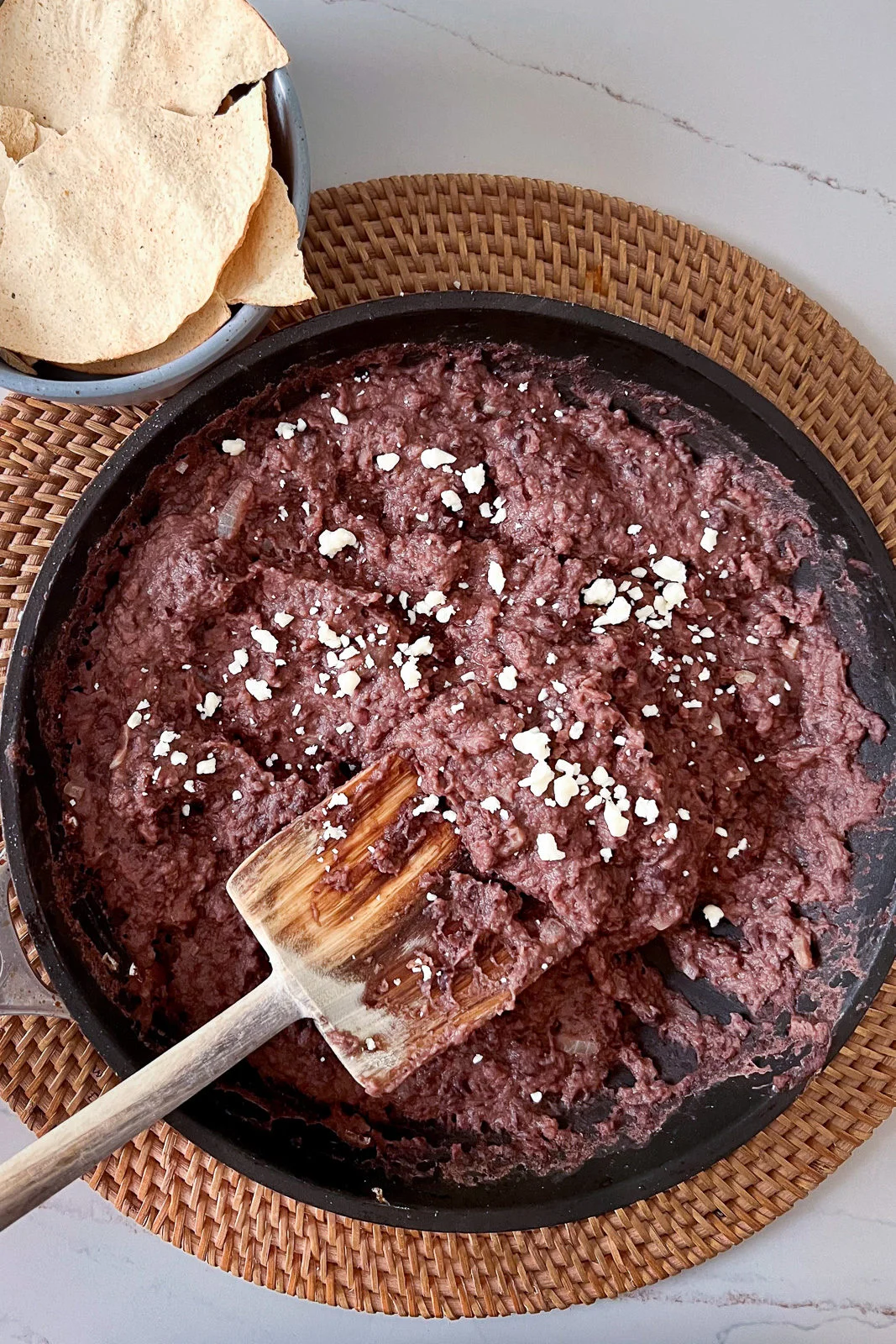 Un sartén con frijoles refritos cremosos hechos en casa, cubiertos con queso blanco desmoronado, sobre un mantel tejido. Una cuchara de madera reposa dentro de los frijoles. En la esquina superior izquierda hay un tazón con totopos crujientes sobre una mesa clara.