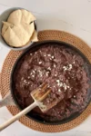 A skillet filled with homemade refried beans sprinkled with cheese sits on a woven placemat. A wooden spatula rests in the pan. Next to it, a gray bowl holds crispy tortilla chips, all on a white marble countertop.