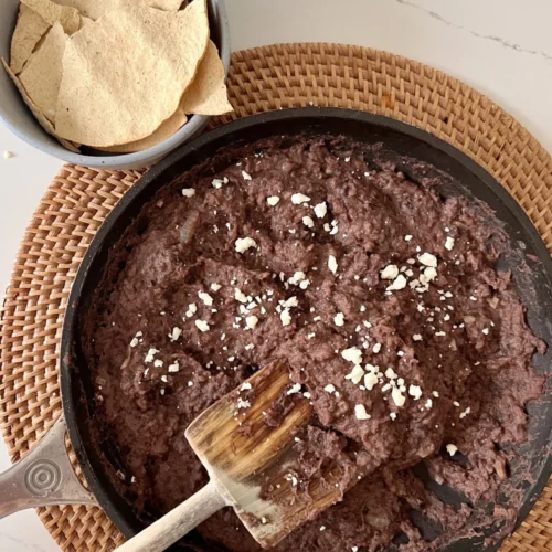 A skillet filled with homemade refried beans sprinkled with cheese sits on a woven placemat. A wooden spatula rests in the pan. Next to it, a gray bowl holds crispy tortilla chips, all on a white marble countertop.