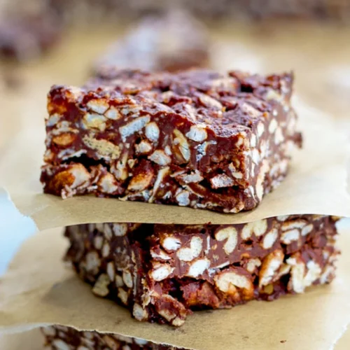 Tres cuadrados de arroz crujiente con chocolate, o barras de chocolate, se apilan sobre papel de pergamino marrón. Rellenas de arroz inflado y recubiertas de chocolate, estas barritas tienen un aspecto texturizado y troceado. El fondo difuminado atrae la atención hacia las tentadoras golosinas.